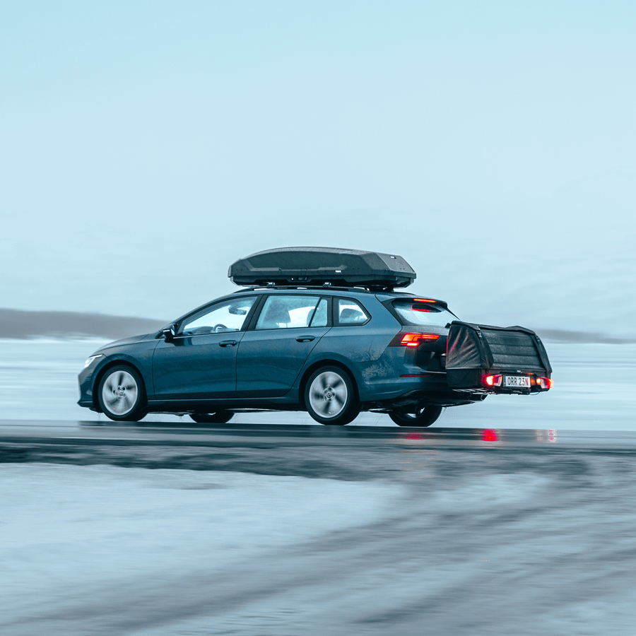 A blue station wagon drives through a snowy landscape with the Thule Onto 2 cargo bag mounted on the towbar and a Thule Force 3 roof box on top, carrying extra gear for winter travel.