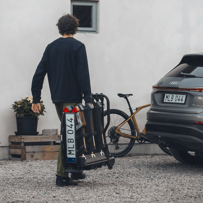 A man in casual outdoor clothing carries a bike rack by its handle, walking between a house and an SUV parked on a gravel driveway.