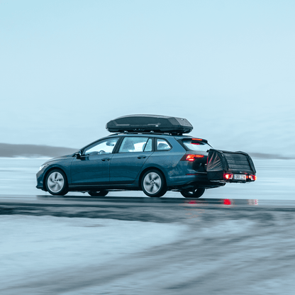 A blue station wagon drives through a snowy landscape with the Thule Onto 2 cargo bag mounted on the towbar and a Thule Force 3 roof box on top, carrying extra gear for winter travel.